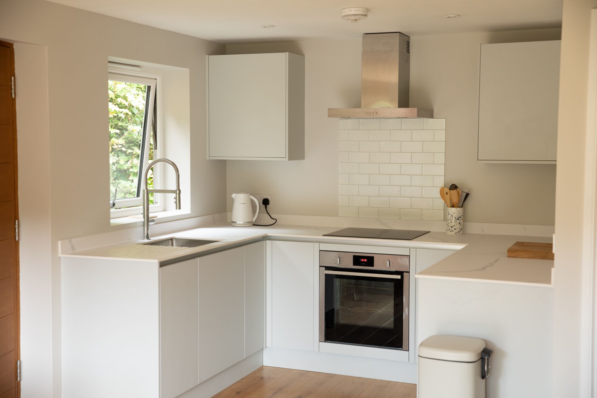 Una cocina moderna con armarios blancos, una campana extractora de acero inoxidable, horno integrado y una ventana con vistas a la vegetación.