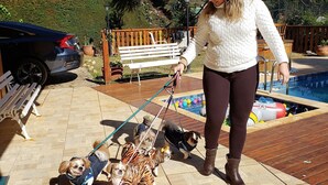A woman walking two small dogs on leashes in a backyard with a pool, a bench, and a car parked nearby. - Suíte Privativa em Jardim1km do Capivari (Campos do Jordão)