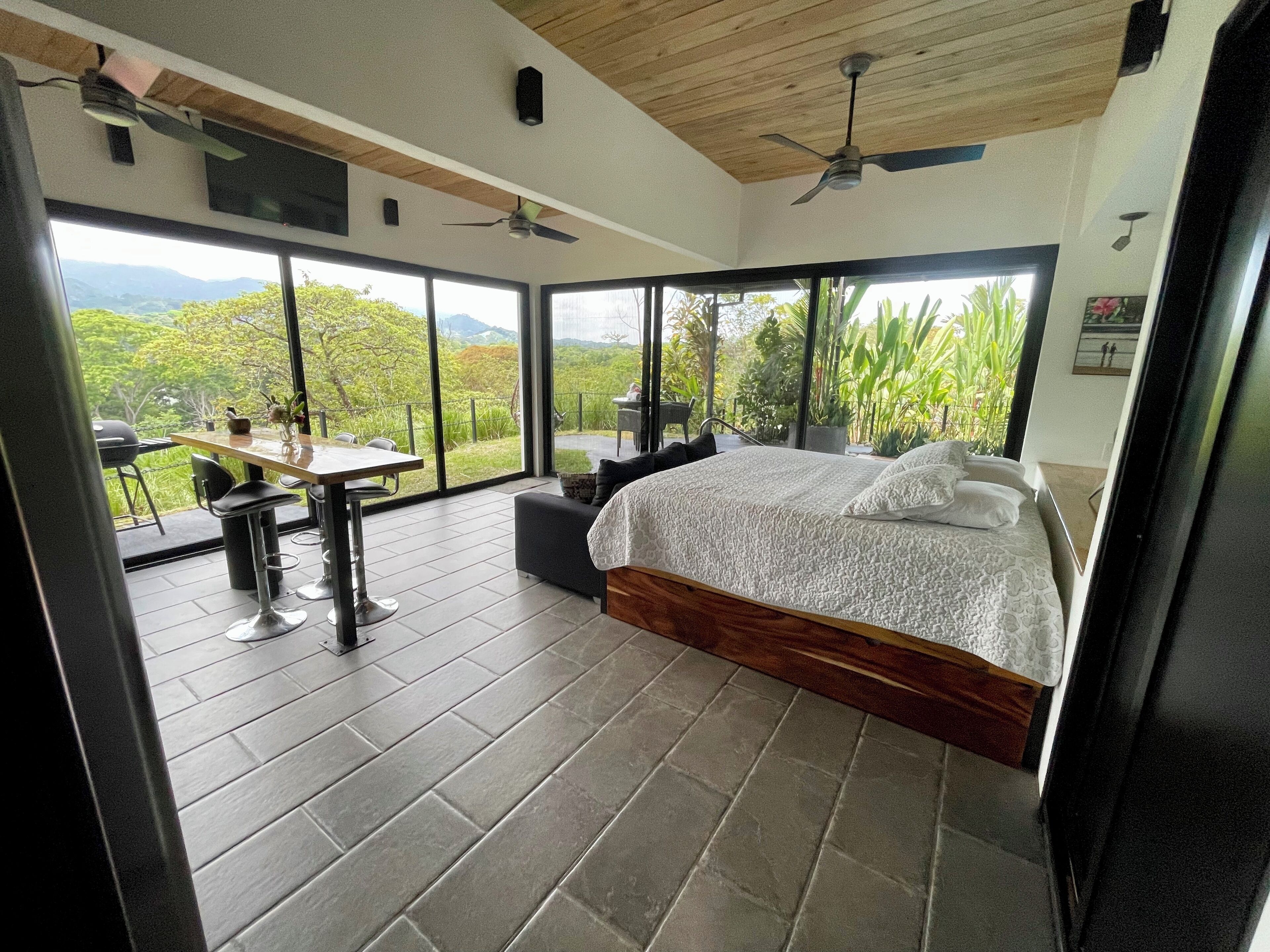 A bedroom with a large bed, a dining table, and chairs, a ceiling fan, and a view of the outdoors.