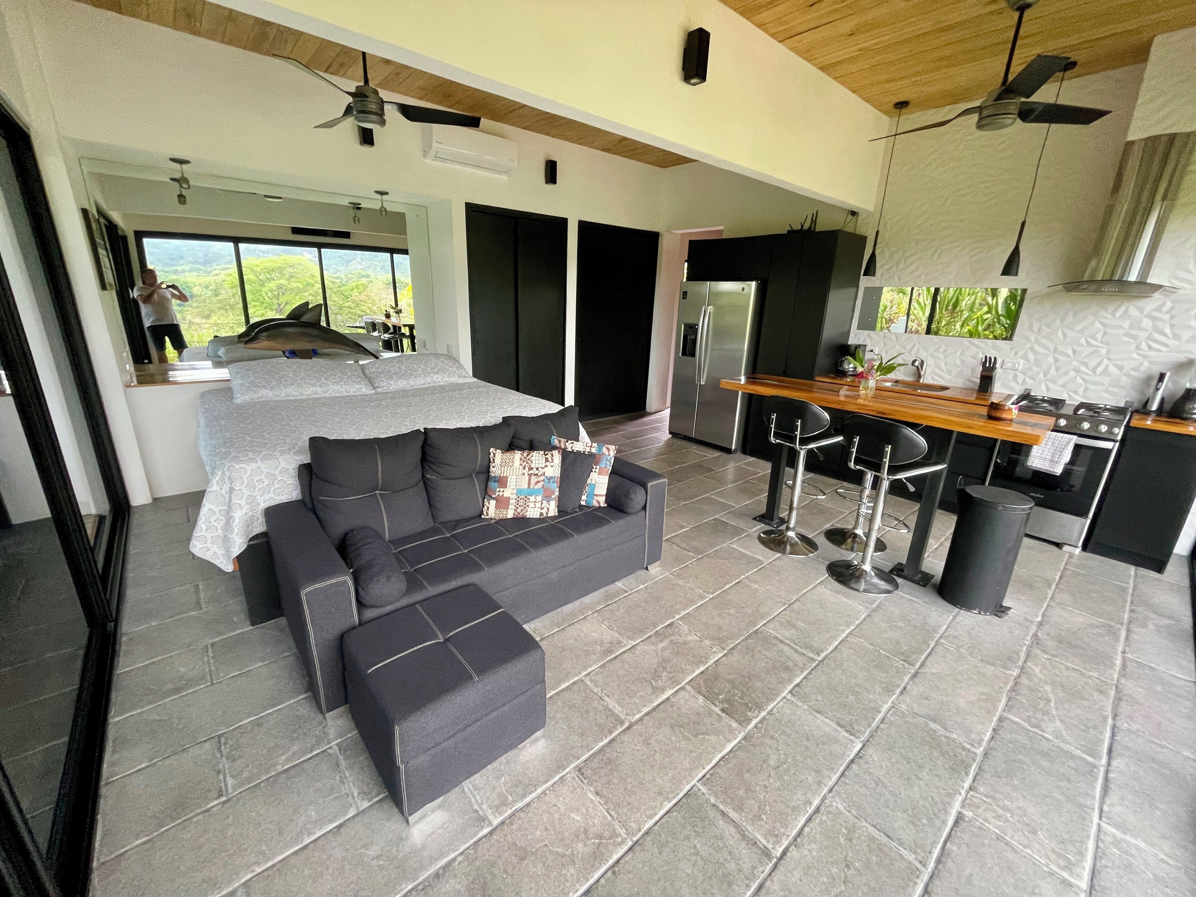 Ein modernes Wohnzimmer mit grauem Sofa, einem Holzesstisch und einer Küchenzeile mit Kühlschrank und Herd.