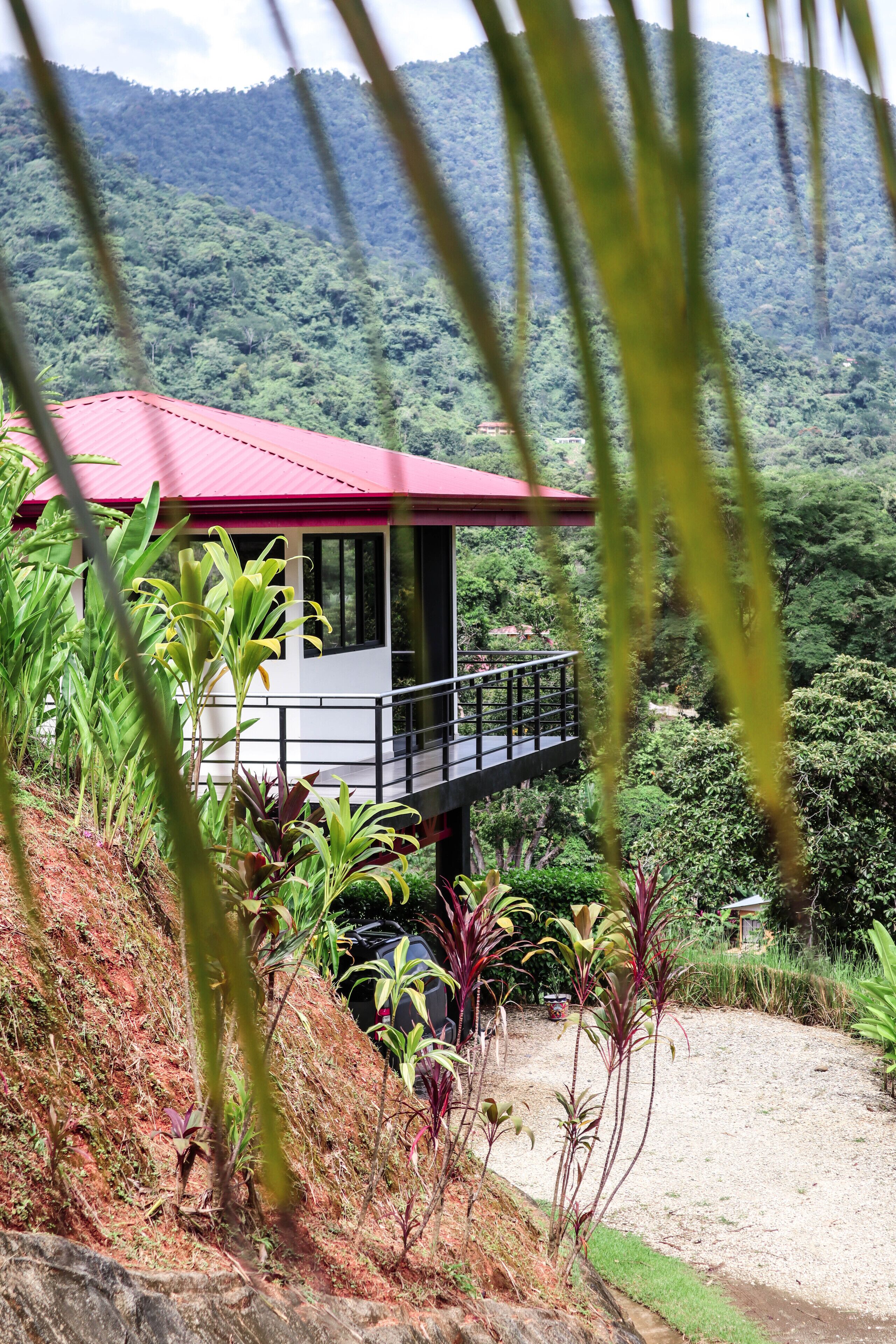 A two-story building with a red roof, surrounded by lush greenery and hills in the background.