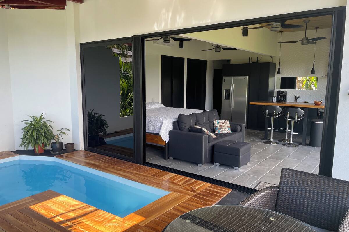 Uma sala de estar moderna com piscina, sofá, mesa de jantar e cozinha visível através de uma porta de vidro.
