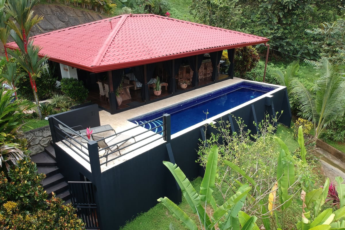 Uma área de piscina moderna ao ar livre, com um pavilhão de telhado vermelho, rodeada por vegetação exuberante.
