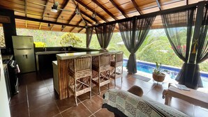 A kitchen with a refrigerator, a sink, and a dining area with a table and chairs. There is a pool visible through the window. - Villa Costa Pura Vida (Ciudad Cortés)