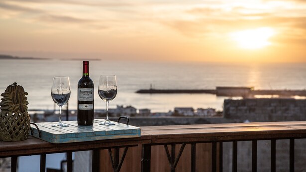 Una mesa con una botella de vino, dos copas y una piña decorativa en una terraza de madera con vistas a la puesta de sol sobre el agua.