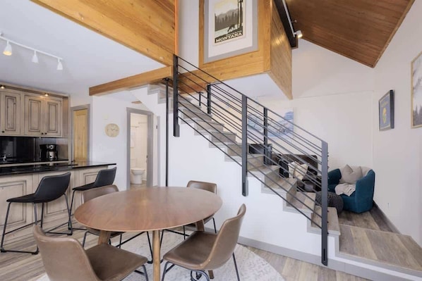 A modern kitchen with wooden beams, a dining area with a round table and chairs, and a staircase with a metal railing.