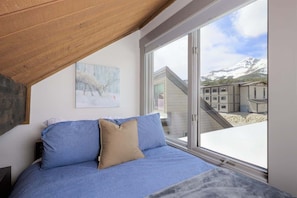 A bedroom with a large window, a bed with blue bedding, a tan pillow, and a painting of sheep on the wall.