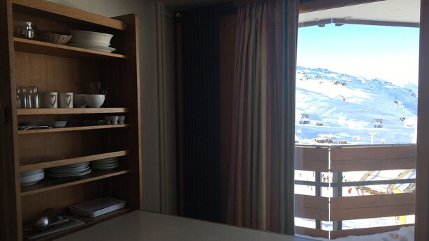Una habitación con una mesa de madera y estantes que contienen platos y tazones. Una ventana con cortinas ofrece vistas a una montaña nevada y un telesilla.