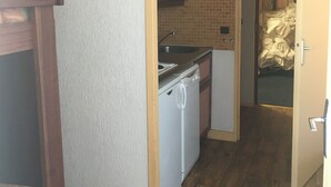 Cocina compacta con lavavajillas, pileta y refrigerador pequeño. Se ve una silla de madera y una cama en la habitación contigua.