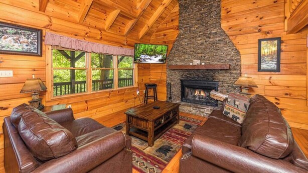 A log cabin interior with a stone fireplace, leather sofas, and a television mounted on the wall. - Pigeon Forge Cabin /Game Room/Dog Friendly/Hot Tub (Pigeon Forge)