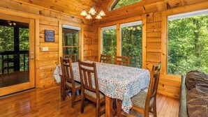 A wooden cabin interior with a dining table set for four, large windows, and a ceiling light fixture.