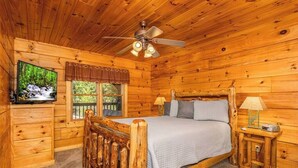 A wooden cabin bedroom with a bed, a television, a window, a ceiling fan, and a nightstand.