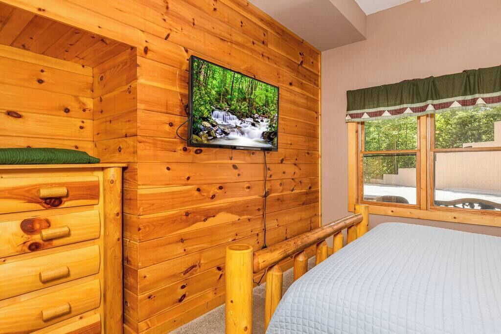A wooden cabin room with a bed, a wooden dresser, a television mounted on the wall, and a window with a view of greenery.