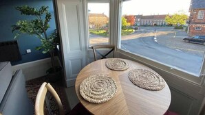 A room with a round wooden table, two woven placemats, a potted plant, and a view of a street with buildings and cars. - Great for exploring North Yorkshire (Easingwold)