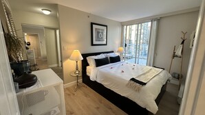 A hotel room with a bed, two bedside lamps, a large window with a view of buildings, a mirror, and a potted plant.
