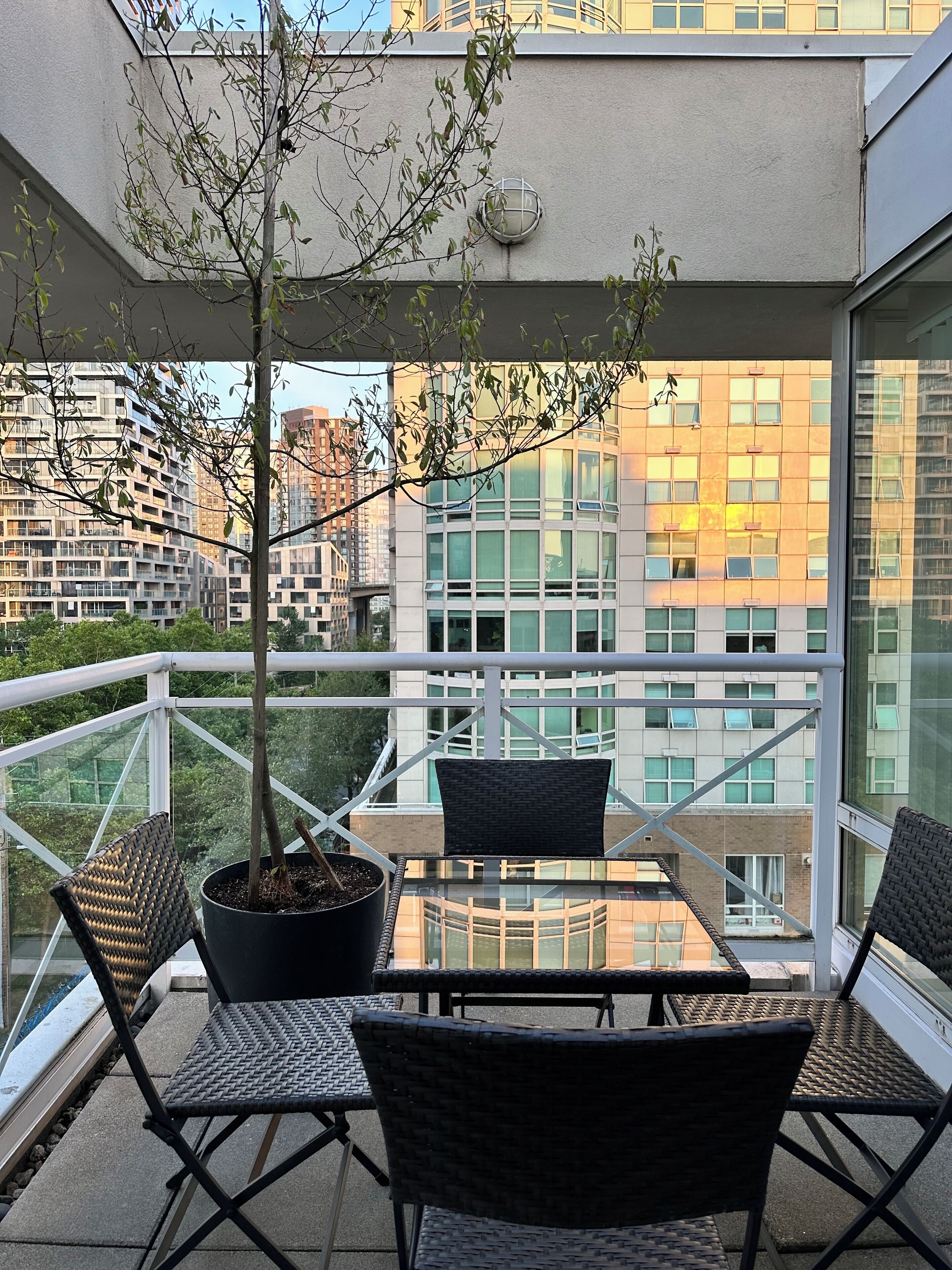 Un balcón con una mesa y sillas de vidrio, un árbol en maceta y vistas a edificios modernos.