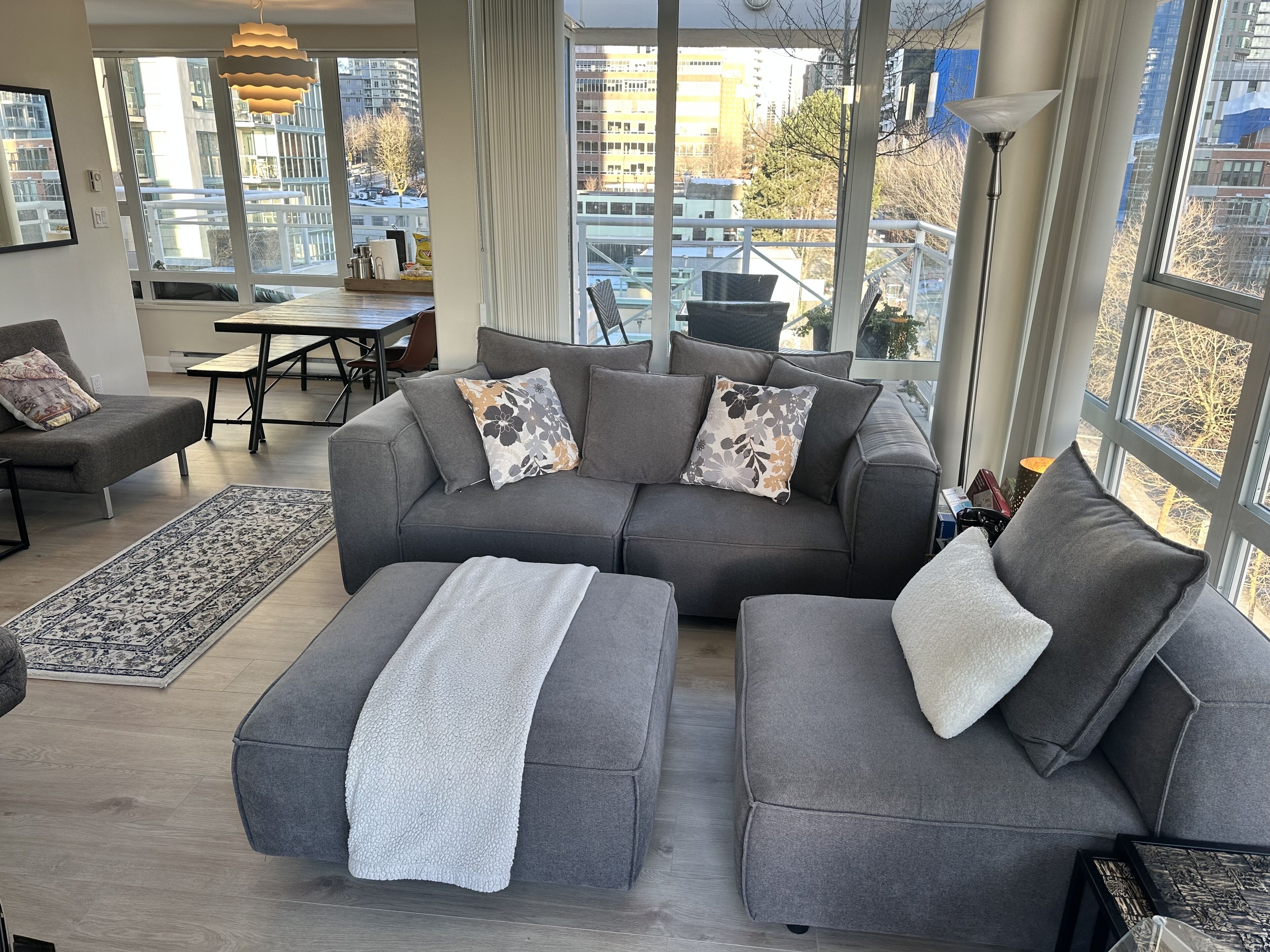 Een moderne woonkamer met een grijze modulaire bank, een kamerkleed met patroon en grote ramen met uitzicht over de stad.