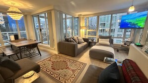 A modern living room with a large TV, comfortable seating, and large windows offering city views.