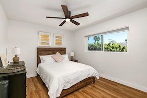 Um quarto com cama de madeira, ventilador de teto, duas obras de arte nas paredes, janela com vista para o verde e mesa de cabeceira com luminária.