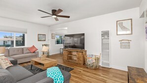 A modern living room with a grey sofa, a wooden coffee table, a TV on a wooden stand, and a ceiling fan. - Renovated 2 BD! Walk to Beach, Restaurants, Bars! (San Diego)