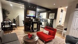 Una sala de estar moderna con un sofá rojo, sillones grises y una mesa de centro. Se observa un área de cocina al fondo con una mesa de comedor y sillas.