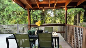 Un patio exterior con una pérgola de madera, luces colgantes, una mesa con sillas y una planta en maceta.