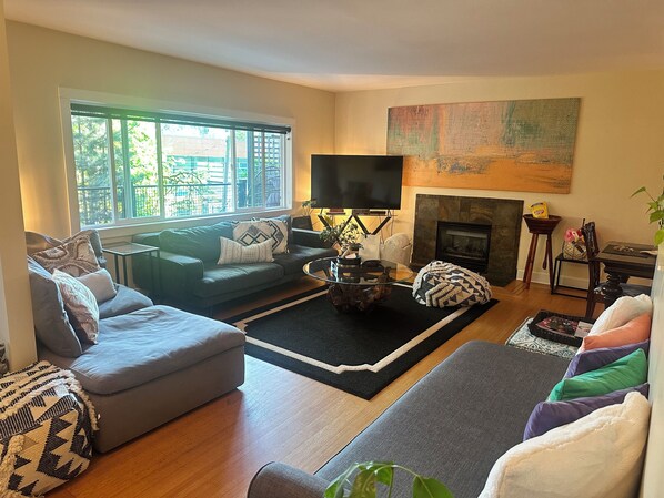 A living room with a sofa, ottoman, coffee table, TV, fireplace, and a large window. - Resort Style Chalet in the Forest with a Tennis Court near Grouse Mountain (North Vancouver)