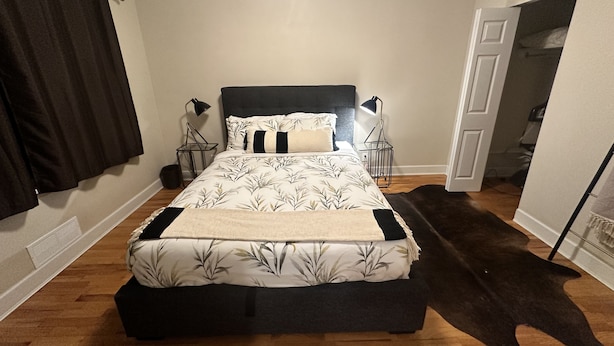 Un dormitorio con una cama, dos mesitas de noche, una alfombra de cuero negro y una puerta que da a otra habitación.