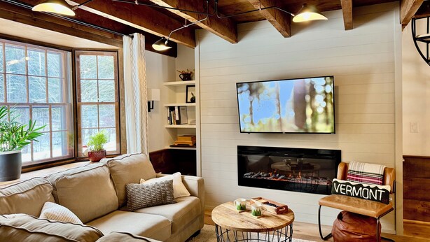 A cozy living room with a sectional sofa, a fireplace, a flat-screen TV, and a bookshelf.