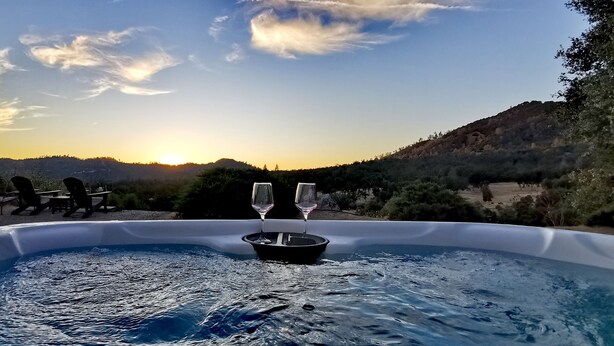 Un jacuzzi con dos copas de vino, rodeado de mobiliario de exterior, con vistas a un paisaje escénico con colinas y árboles al atardecer.