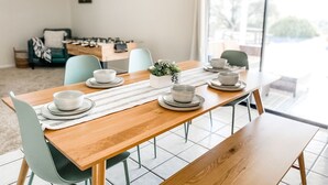 Un comedor con una mesa de madera dispuesta para cuatro personas, rodeada de sillas. Se ve una cocina al fondo y un letrero en la pared dice 'GAME ON'.