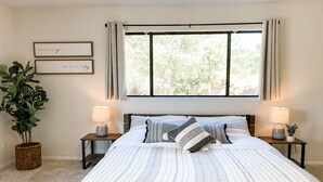 Un dormitorio con una cama grande, dos mesitas de noche con lámparas, una planta en maceta y una ventana con cortinas.