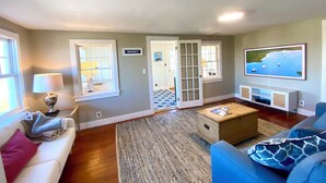 A living room with a white couch, blue sofa, and a coffee table. There is a TV on a white stand, a lamp on a side table, and a door labeled "MADAKET".