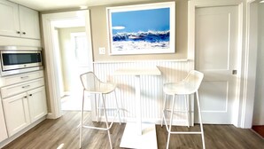 A kitchen with white cabinets, a microwave, two white bar stools, and a framed picture of a beach scene.