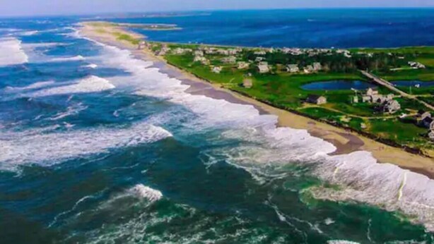 An aerial view of a coastal area with a beach, green fields, and a few buildings.