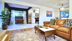 Una sala de estar con un sofá de cuero marrón, una mesa de centro de madera y un tapete estampado. Se observa un comedor con una mesa y sillas de madera, y una cocina visible a través de un arco abierto.