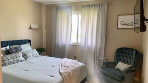 Ein Schlafzimmer mit Bett, Sessel, Nachttisch und einem Fenster mit Vorhängen.