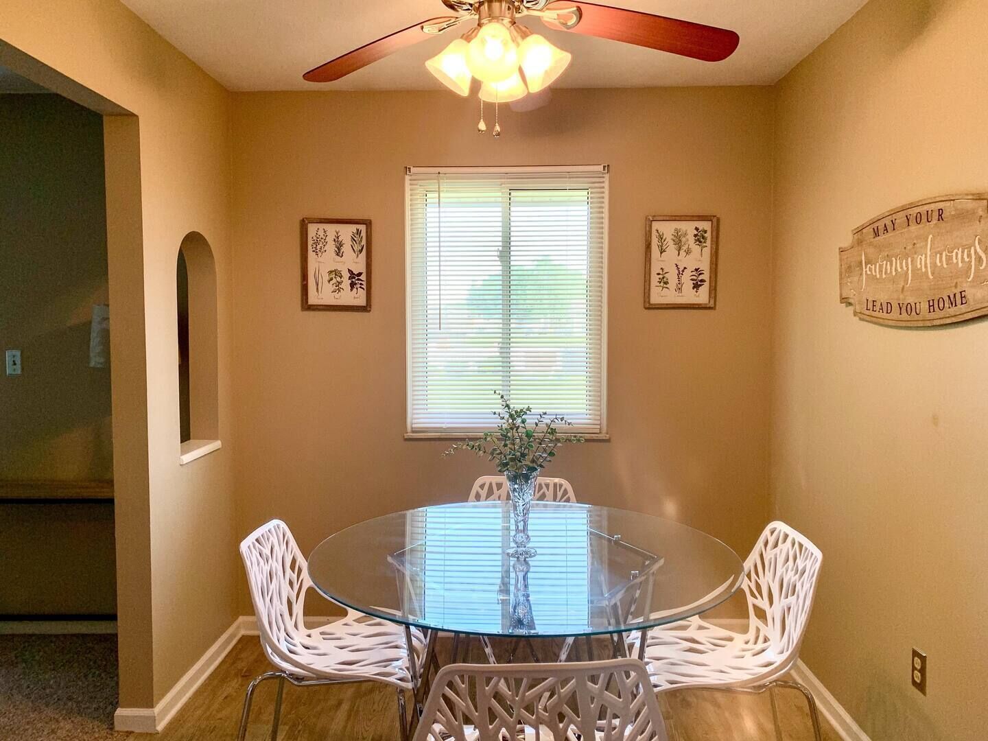 A dining area with a glass table and white chairs, a ceiling fan, and framed artwork on the walls.