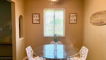 A dining area with a glass table and white chairs, a ceiling fan, and framed artwork on the walls.