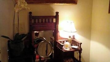 Una habitación con una silla de madera, una mesita con una lámpara, un cuadro enmarcado y un jarrón con flores.