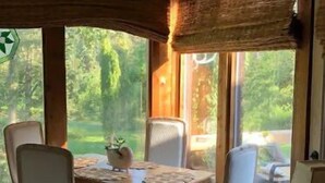 Un espace repas avec une table et des chaises en bois, de grandes fenêtres donnant sur un paysage verdoyant et un décor intérieur de style rustique.