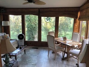 Een kamer met een eettafel en stoelen, een plafondventilator en grote ramen met uitzicht op groen.