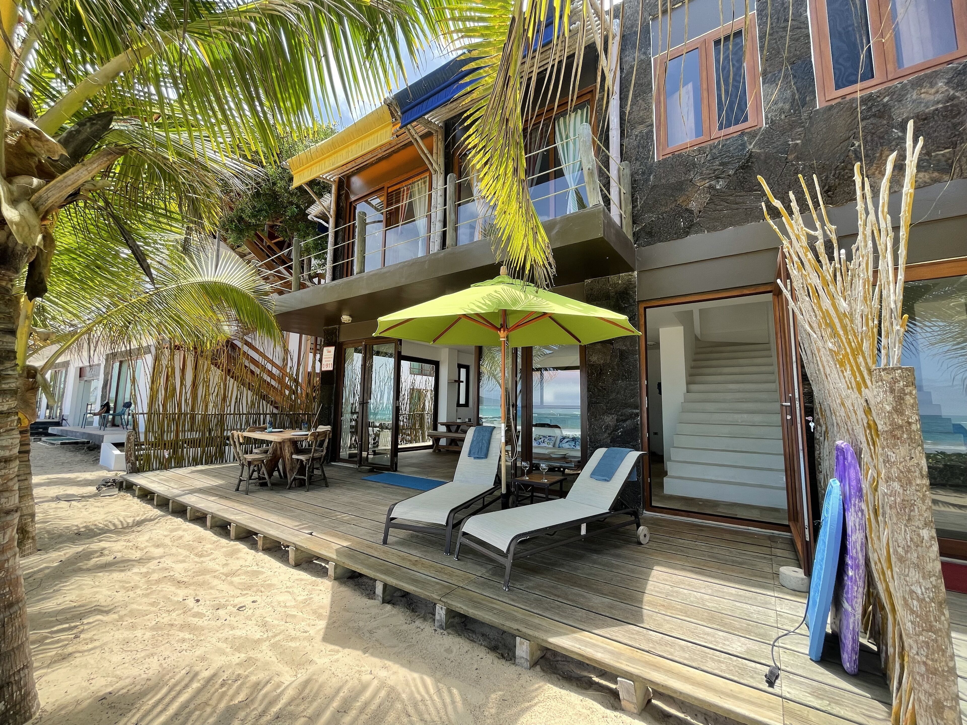 Ein Strandgrundstück mit Holzterrasse, Liegestühlen und einer überdachten Sitzgruppe mit Sonnenschirmen.