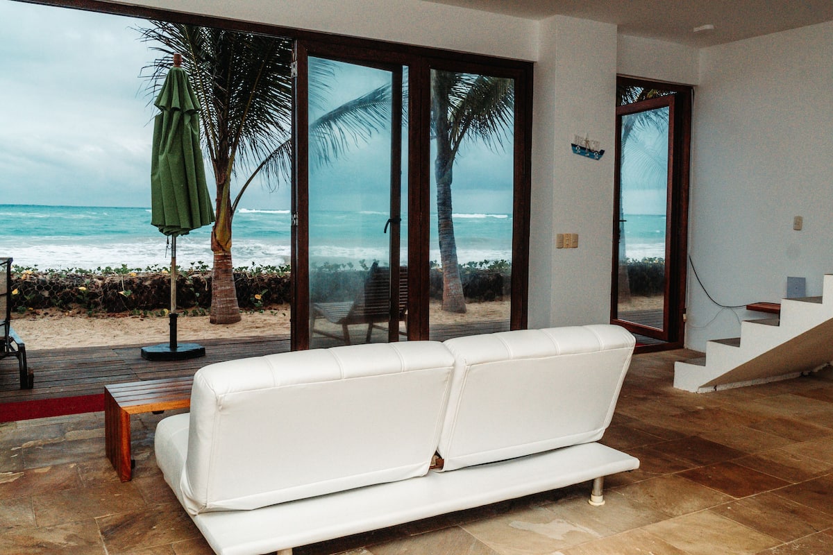 A modern living room with a white sofa, a wooden bench, and a view of the ocean through large glass doors.