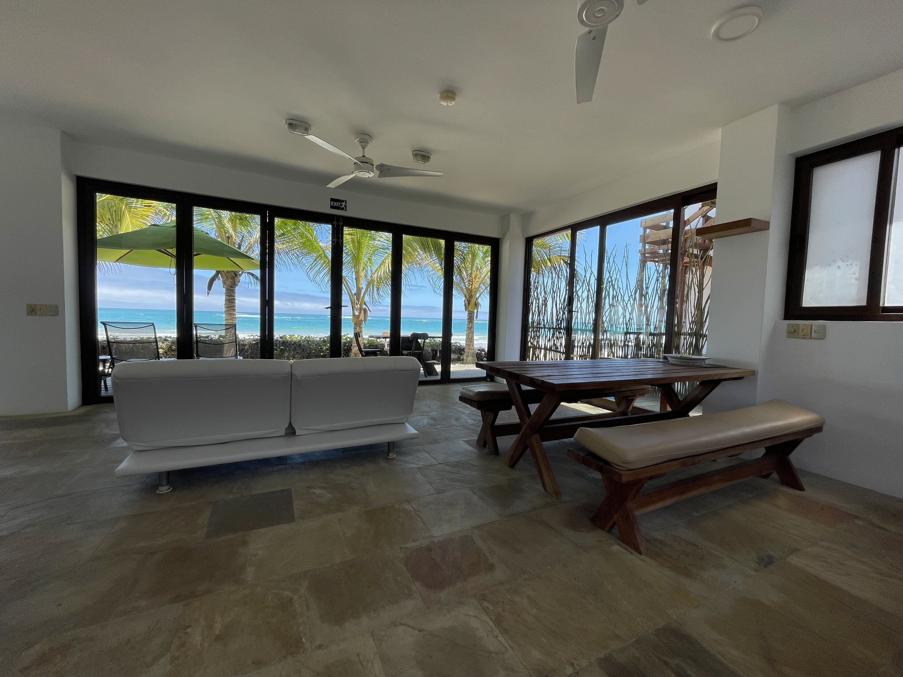 A spacious room with a large window overlooking a beach, a white sofa, a wooden table, and a ceiling fan.