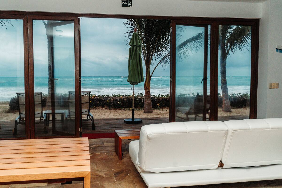 A room with a white sofa, wooden bench, and a view of the beach through large glass doors.