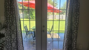 A view through a glass door with a geometric patterned curtain, leading to an outdoor patio with a red umbrella and green chairs.