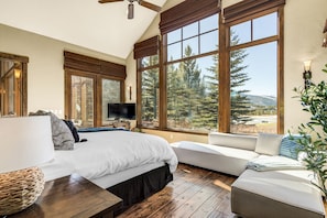 A bedroom with a large bed, a television, and a view of trees and mountains through large windows.
