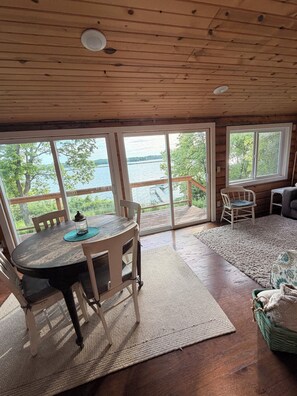Interior de una cabaña de madera con una mesa de comedor, sillas, un tapete, un sofá y ventanas grandes con vistas a un lago.