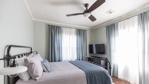 Une chambre à coucher comprenant un lit, une table de chevet, une lampe, un ventilateur de plafond, une télévision et de grandes fenêtres avec des rideaux.